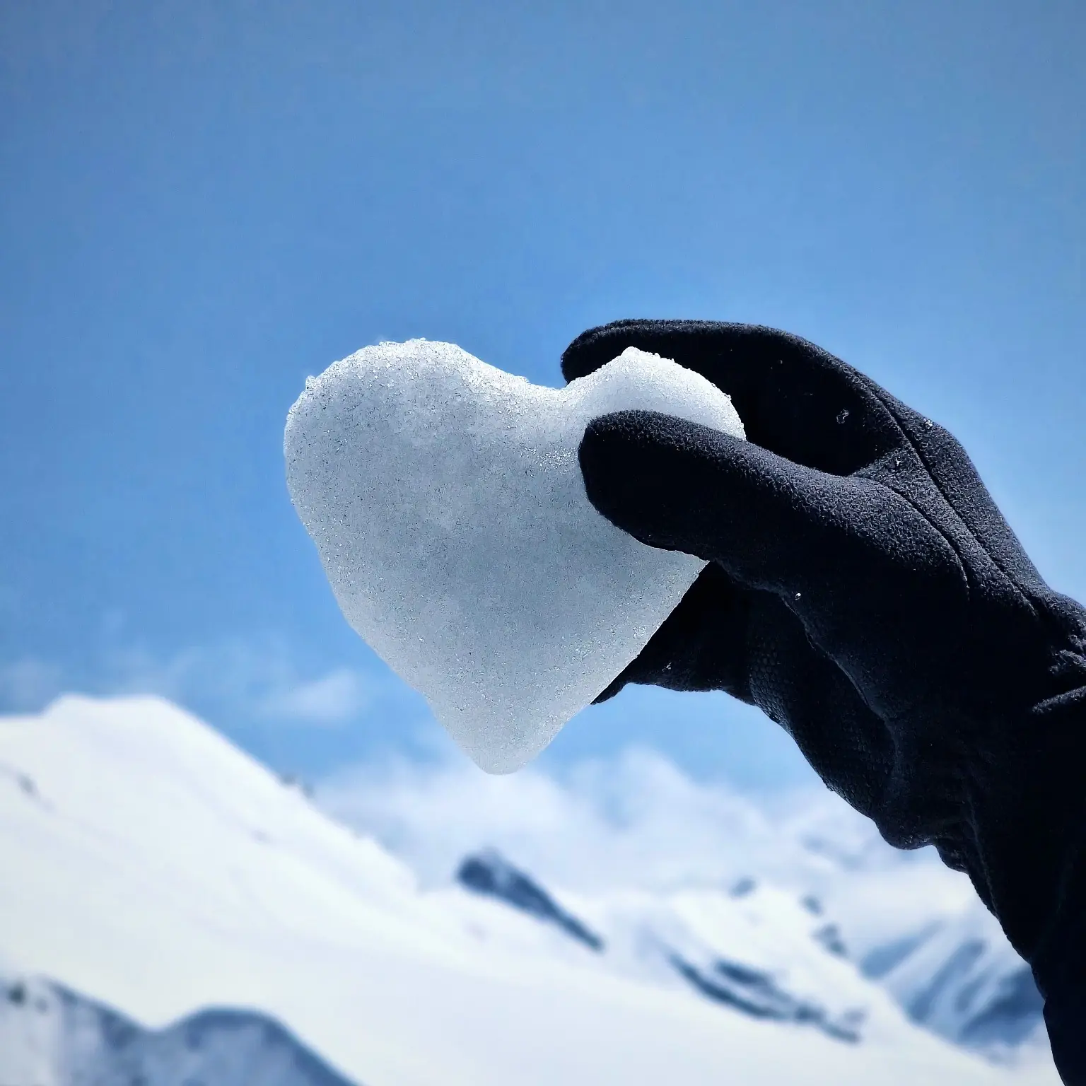 Black-gloved hand holding a heart-shaped piece of ice against snowy mountains