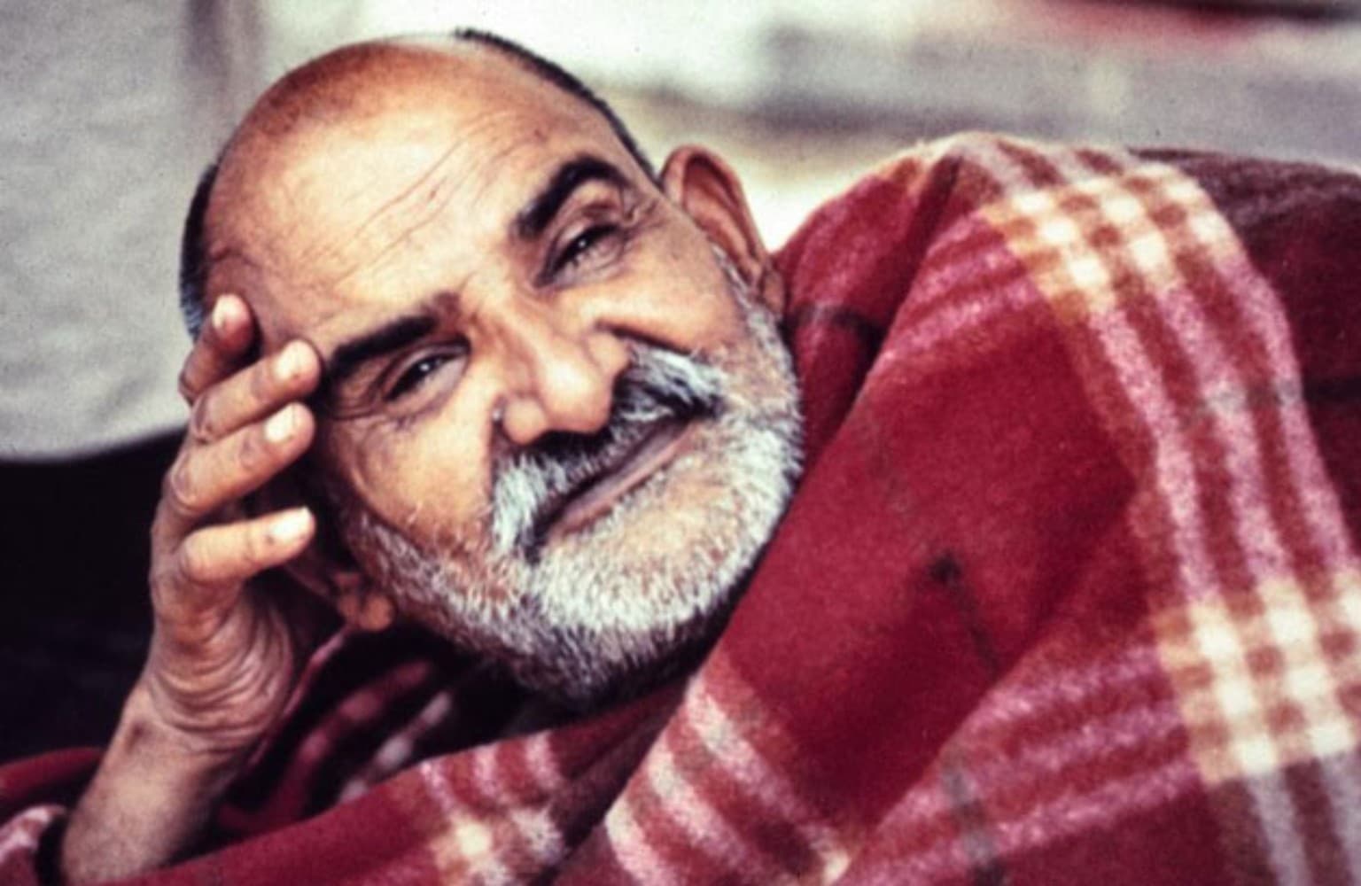 Maharajji smiling, resting his head on his hand and wrapped in a red checked blanket