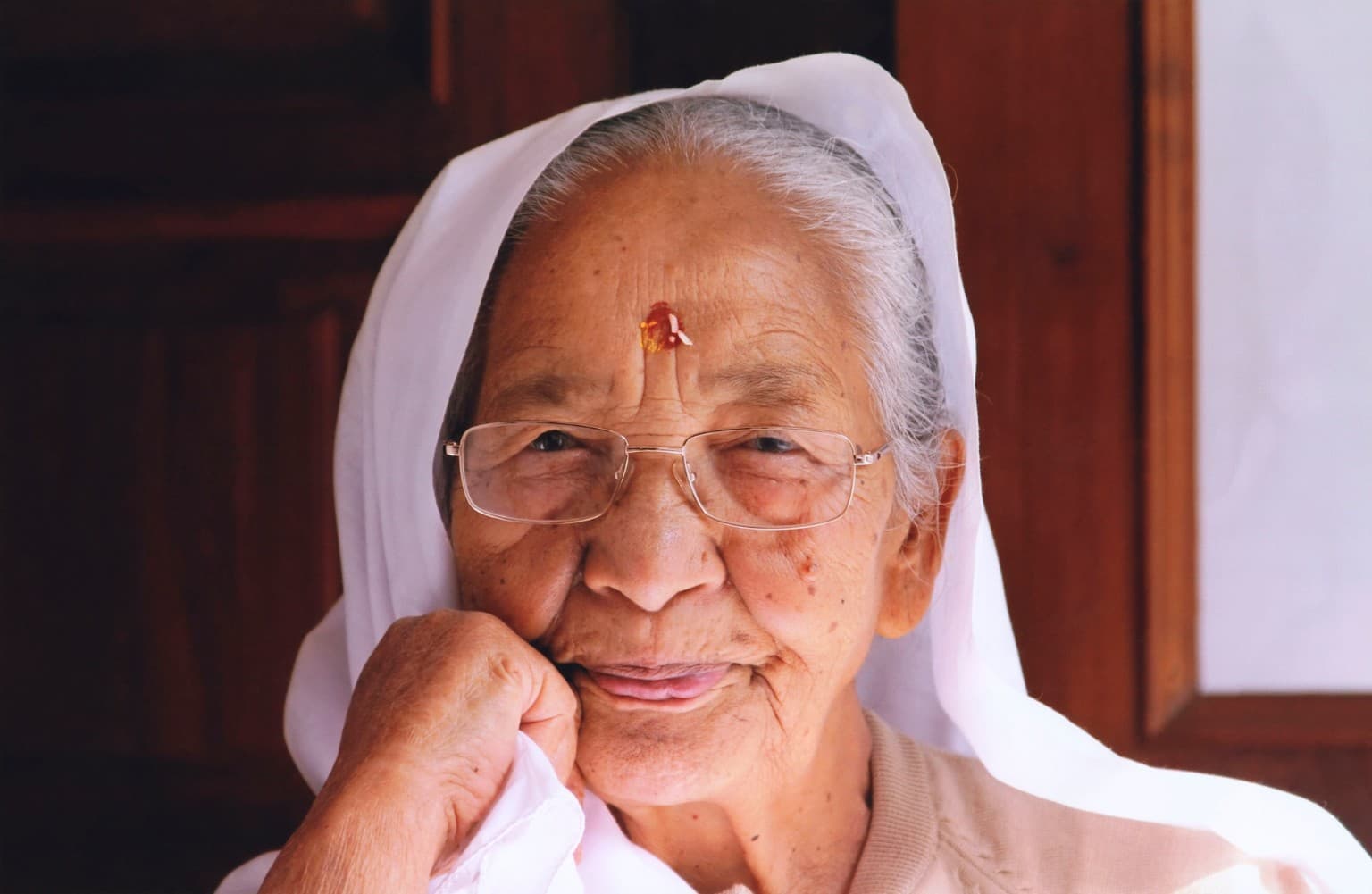 Sri Siddhi Ma smiling, wearing glasses and a white head covering, with her hand resting against her cheek